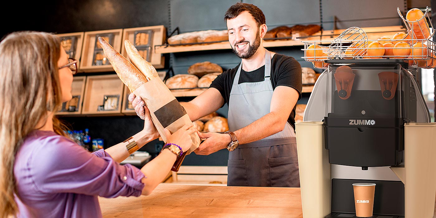 un venditore del panificio porge delle baguette appena sfornate ad una cliente, in primo piano si intravede un spremiagrumi Zummo sfocato